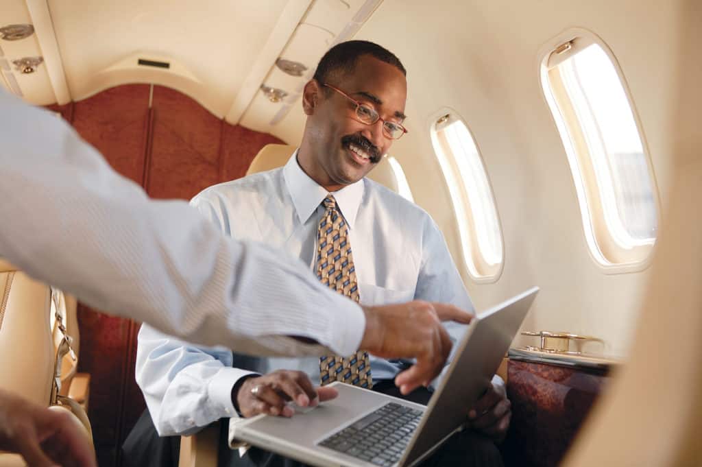 A man pointing at a laptop screen while the other scrolls through it while being seated on a plane